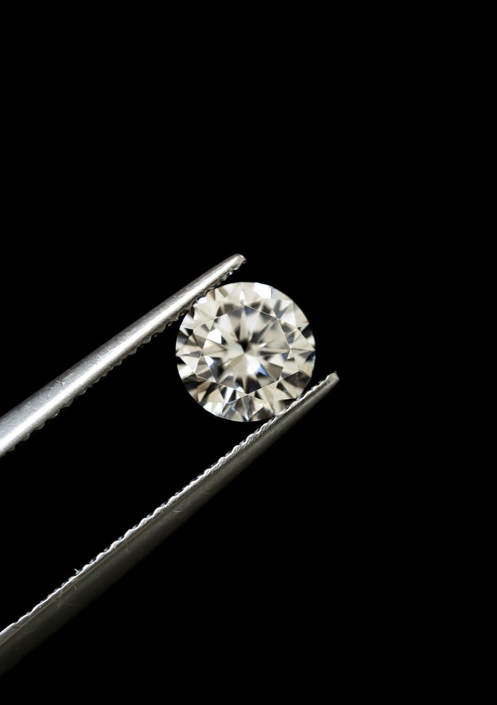 Round diamond gemstone held in tweezers for inspection, minimalist black backdrop
