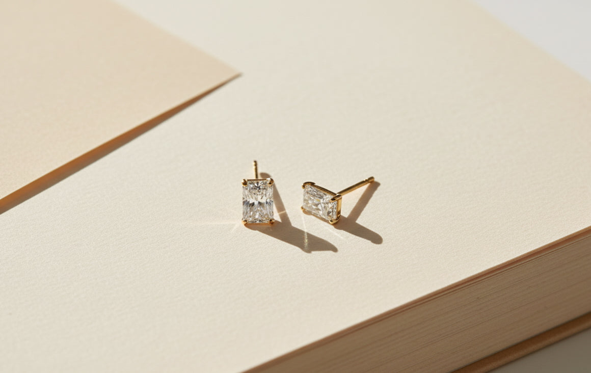 pair of emerald-cut lab-grown diamond stud earrings in 18k gold resting on a sunlit beige book